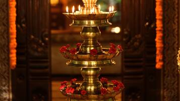 A traditional brass lamp with multiple tiers, adorned with red flowers, placed in a richly decorated room with ornate wooden paneling and a mirror reflecting the interior.