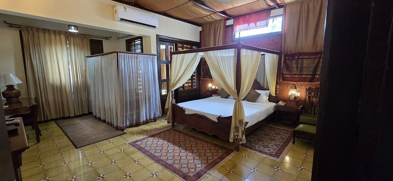 A room with a four-poster bed, curtains, a desk with a lamp, and a tiled floor.