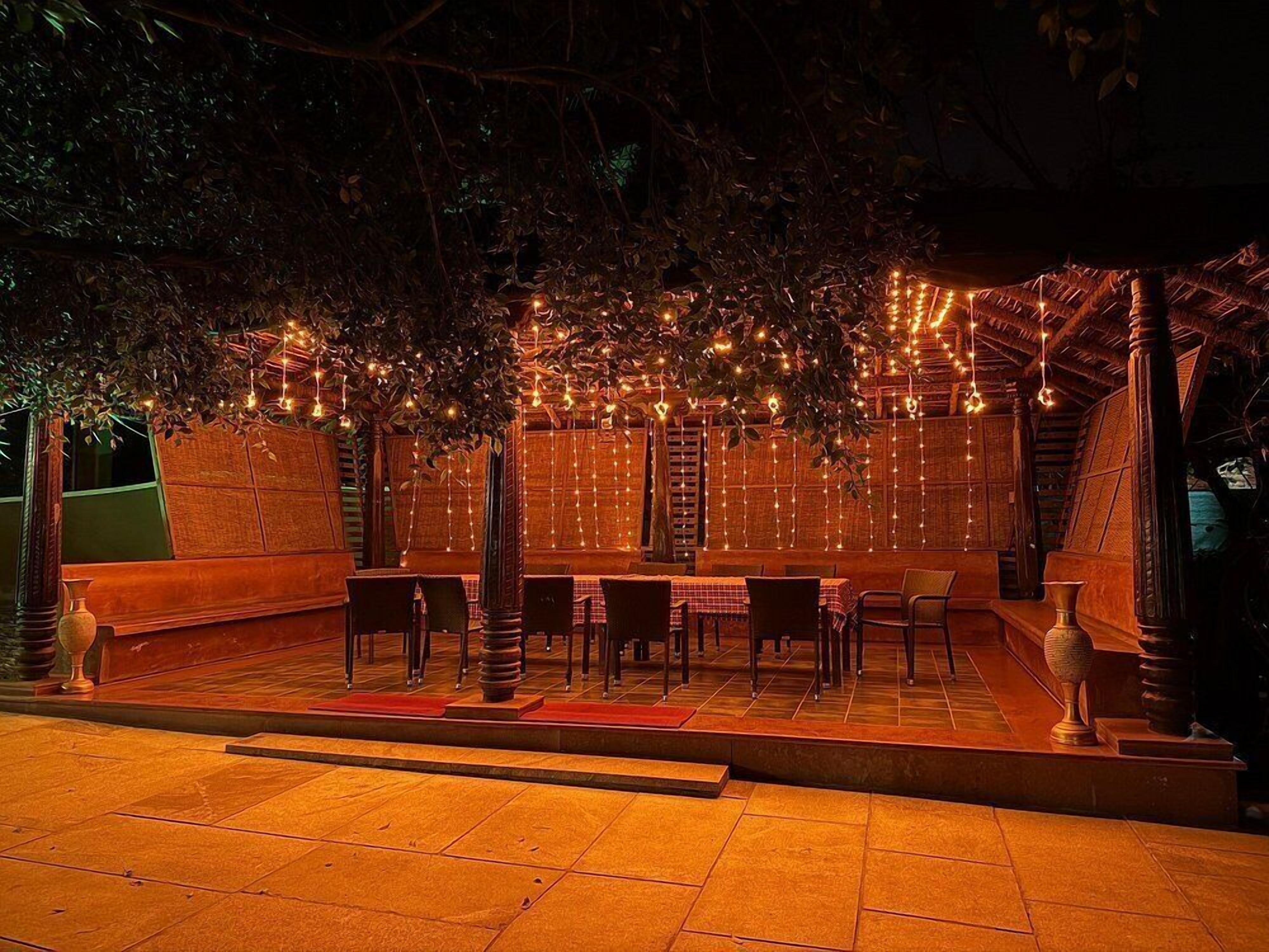 Zona pranzo all'aperto con tavoli e sedie, circondata da alberi e un muro in legno con luci a sospensione.