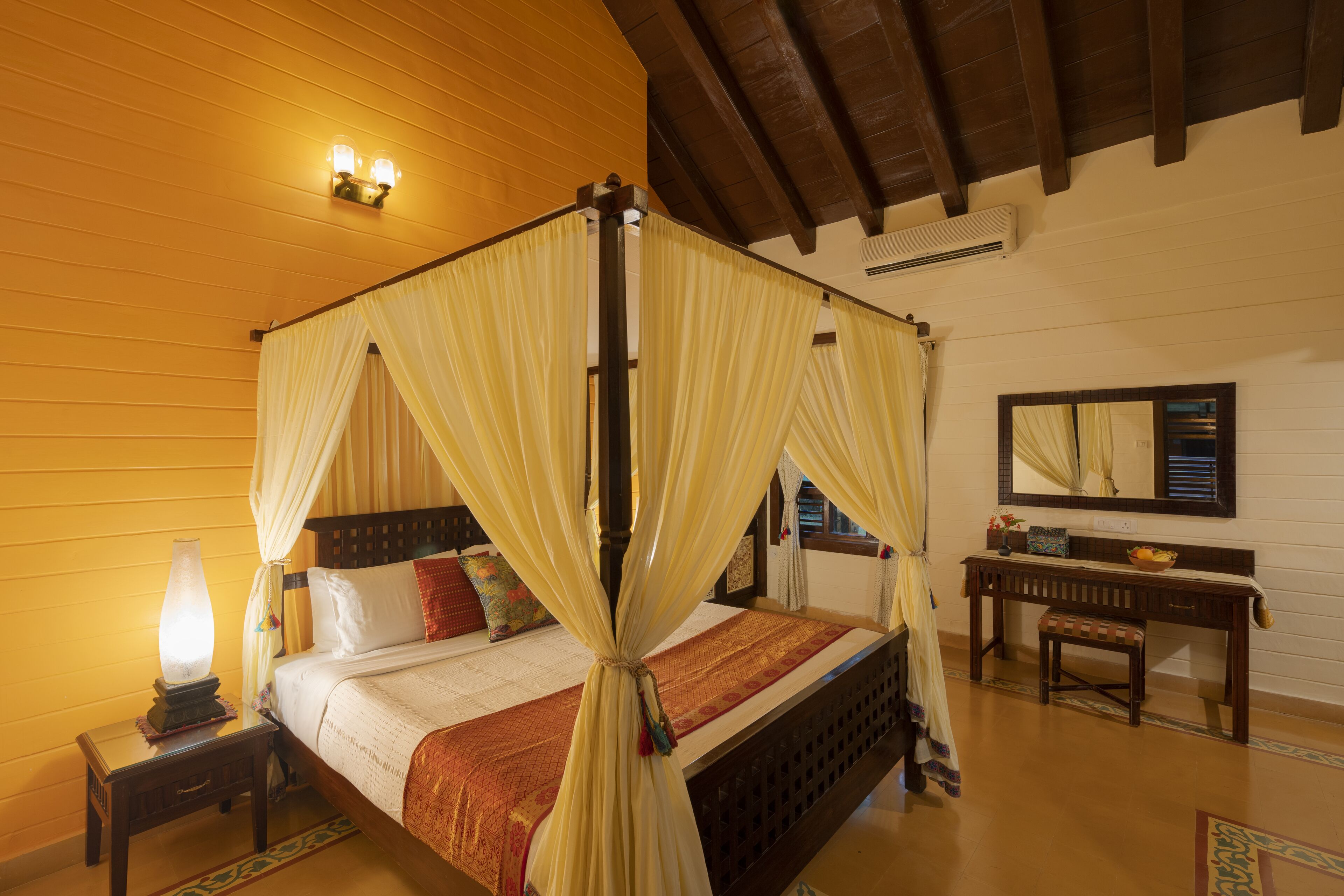 A four-poster bed with draped curtains, a bedside table with a lamp, a wooden bench, and a mirror on the wall.