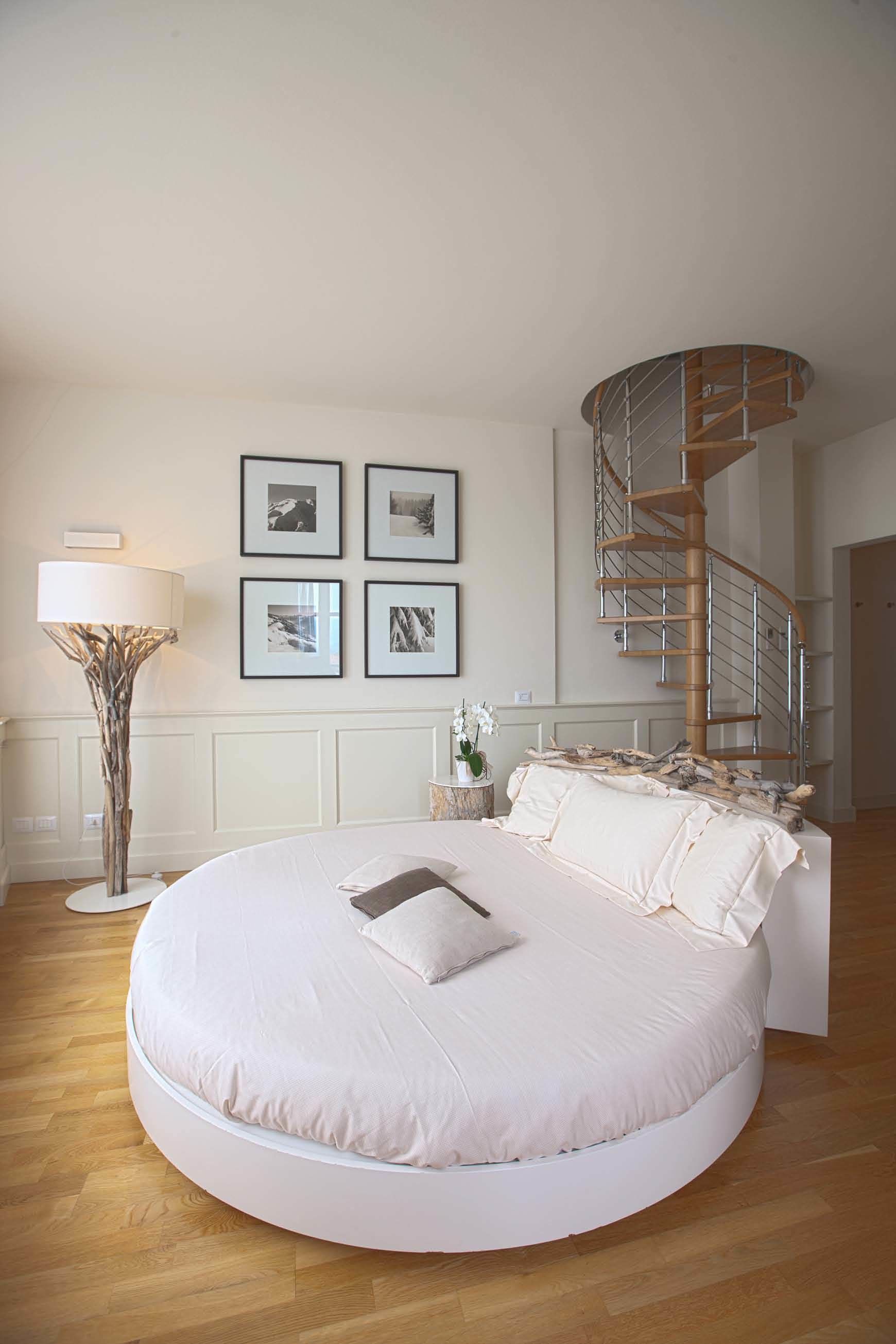 A circular bed with a white headboard, a spiral staircase, and framed pictures on the wall.