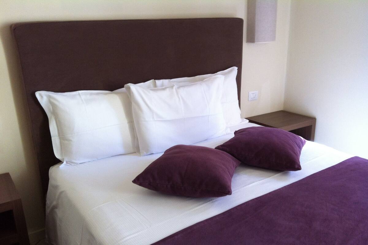 A bed with white bedding and two purple pillows, a brown headboard, and a small wooden nightstand.
