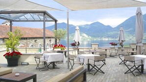 Una terraza al aire libre con mesas y sillas, un área de estar cubierta y vistas a montañas y un lago.