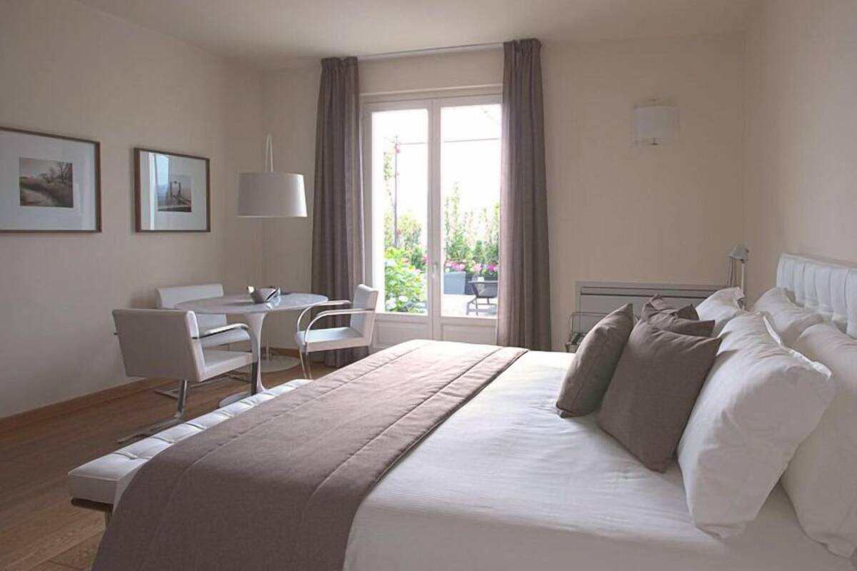A modern hotel room with a large bed, a desk, and a chair. There is a window with curtains and a view of greenery outside.