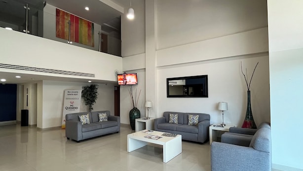 Um hall amplo com dois sofás cinzentos, uma mesa de centro branca e uma TV fixada na parede.