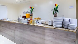 Uma área de recepção com balcão de madeira, quadro branco, máquina de café e dois vasos com plantas e flores vermelhas e amarelas.