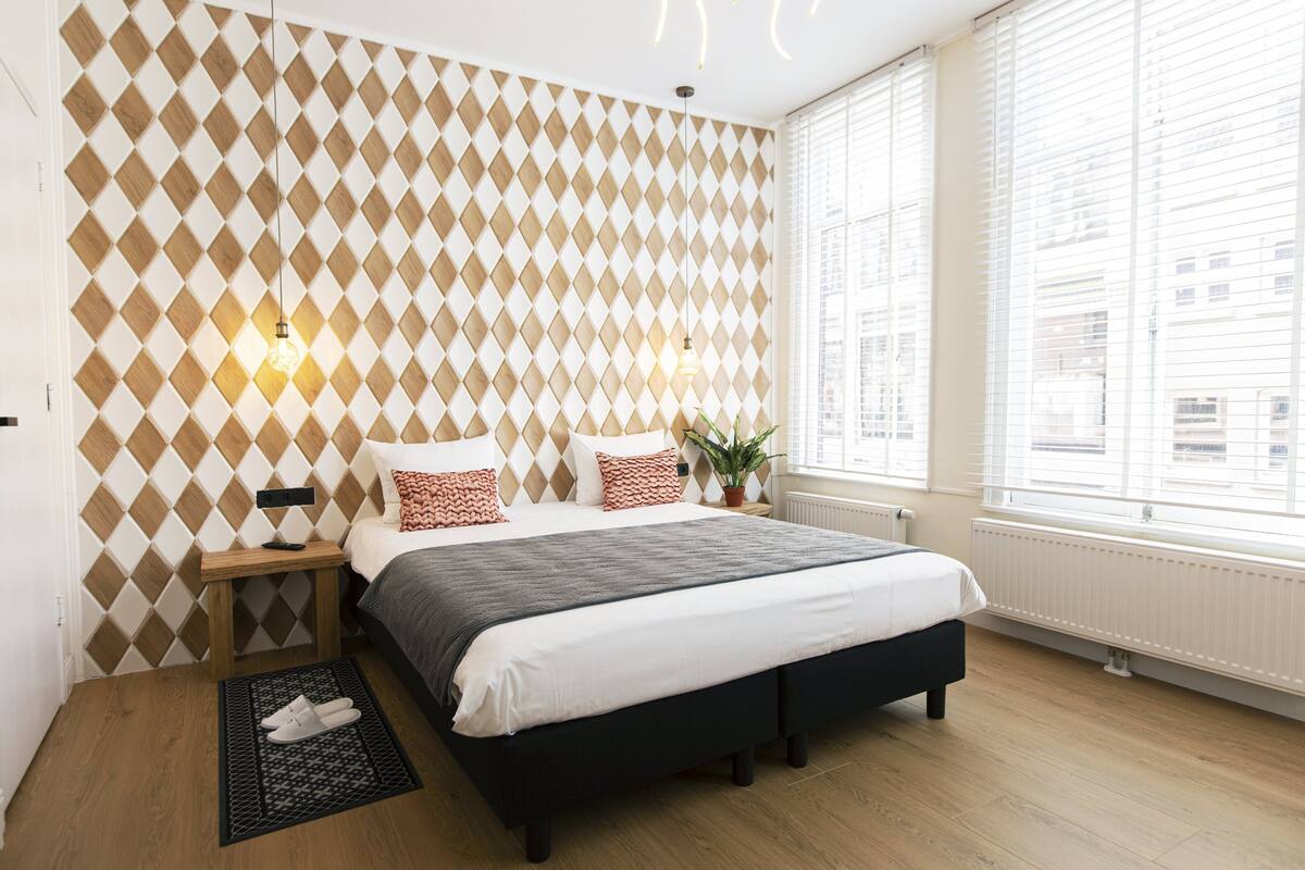 A modern bedroom with a geometric patterned wall, a large bed with a dark headboard, bedside tables with lamps, and a view of the city through large windows.
