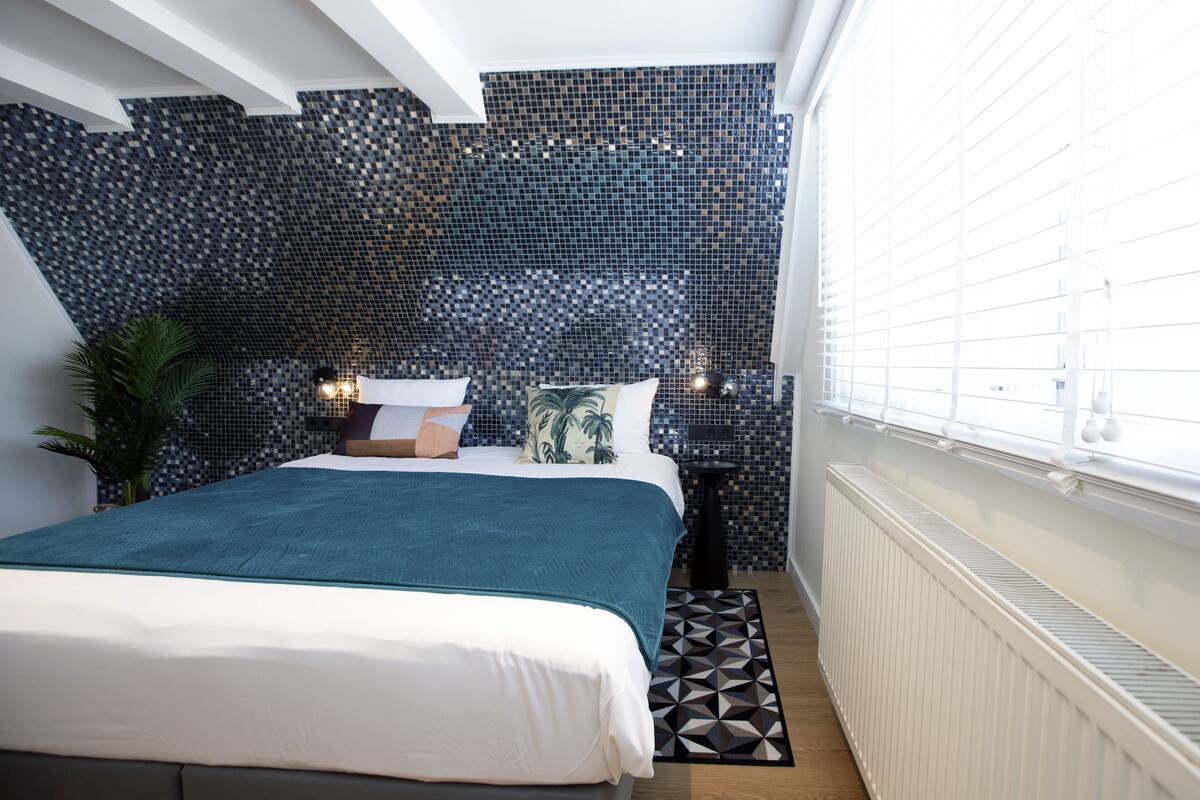 A bedroom with a bed, a nightstand, a potted plant, and a window with blinds.