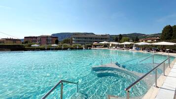 Une grande piscine extérieure à l’eau claire, entourée de chaises longues et de parasols, avec des bâtiments et des collines en arrière-plan.