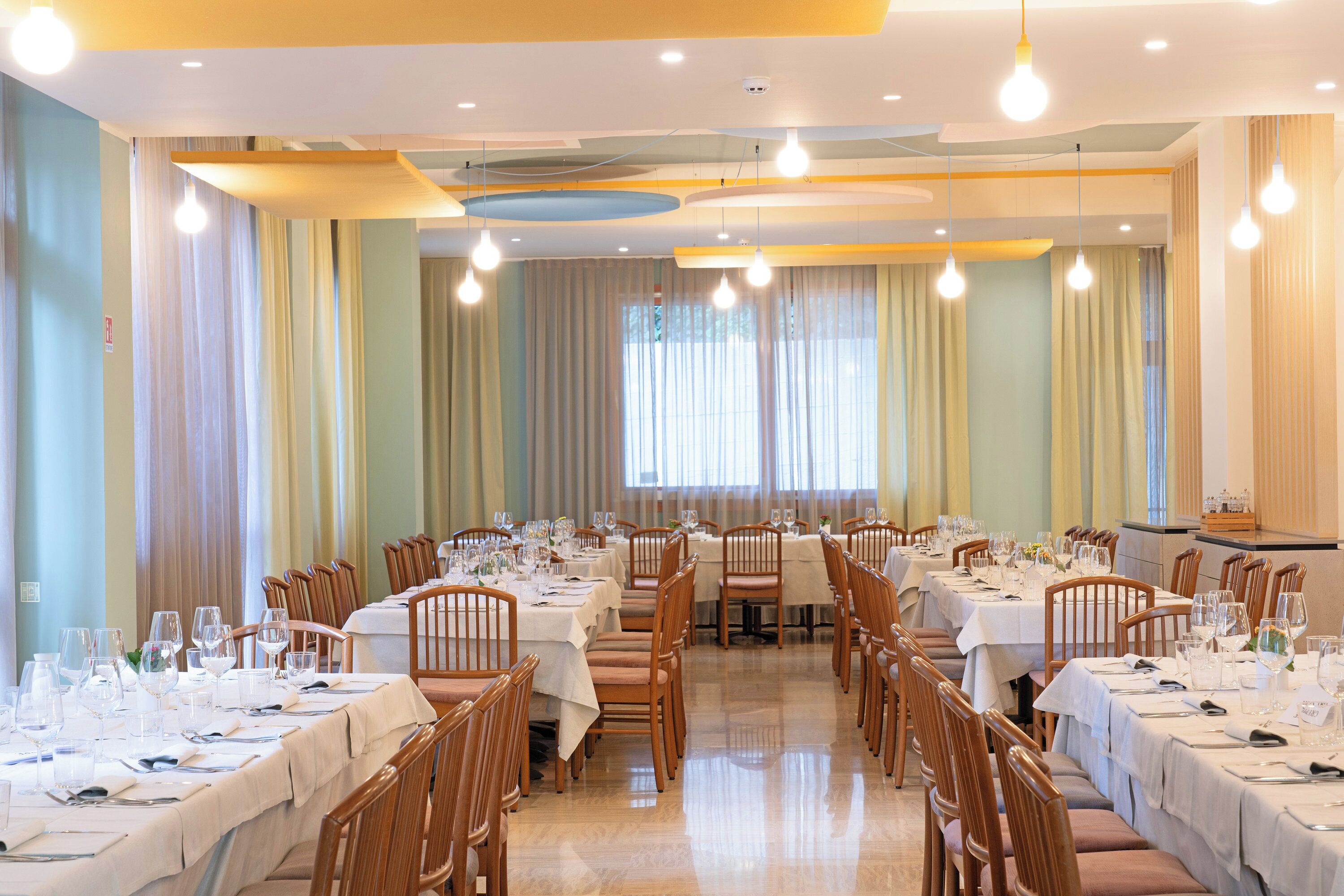 A dining area with multiple tables set for service, wooden chairs, and a ceiling with hanging light fixtures.