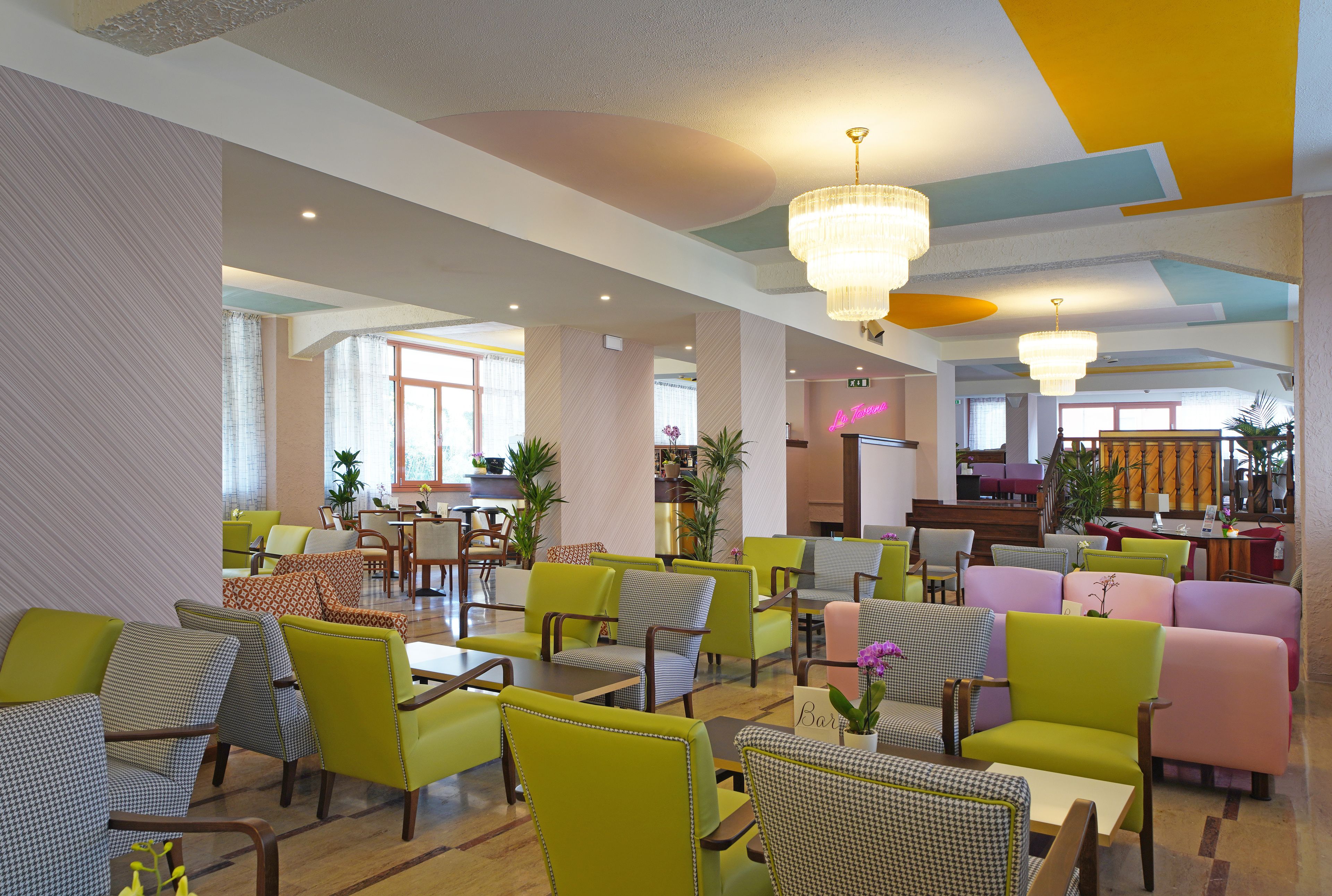 A hotel lobby with multiple seating areas, including green and pink chairs, and a reception desk in the background.