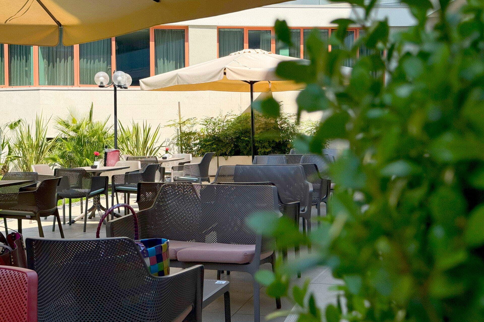 Een terras met tafels en stoelen, een grote parasol, en een gebouw op de achtergrond.
