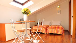 Una habitación con tragaluz, una mesa de comedor con sillas, una cama con ropa de cama escocesa y un jarrón blanco con flores.