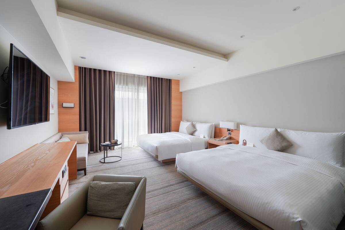A hotel room with two beds, a desk, a chair, a TV, and a window with curtains.