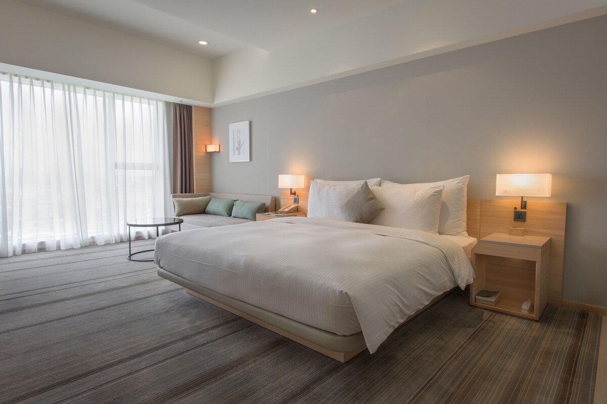 Een moderne hotelkamer met een groot bed, bedlampen, een kleintje tafeltje en uitzicht op het landschap door dunne gordijnen.