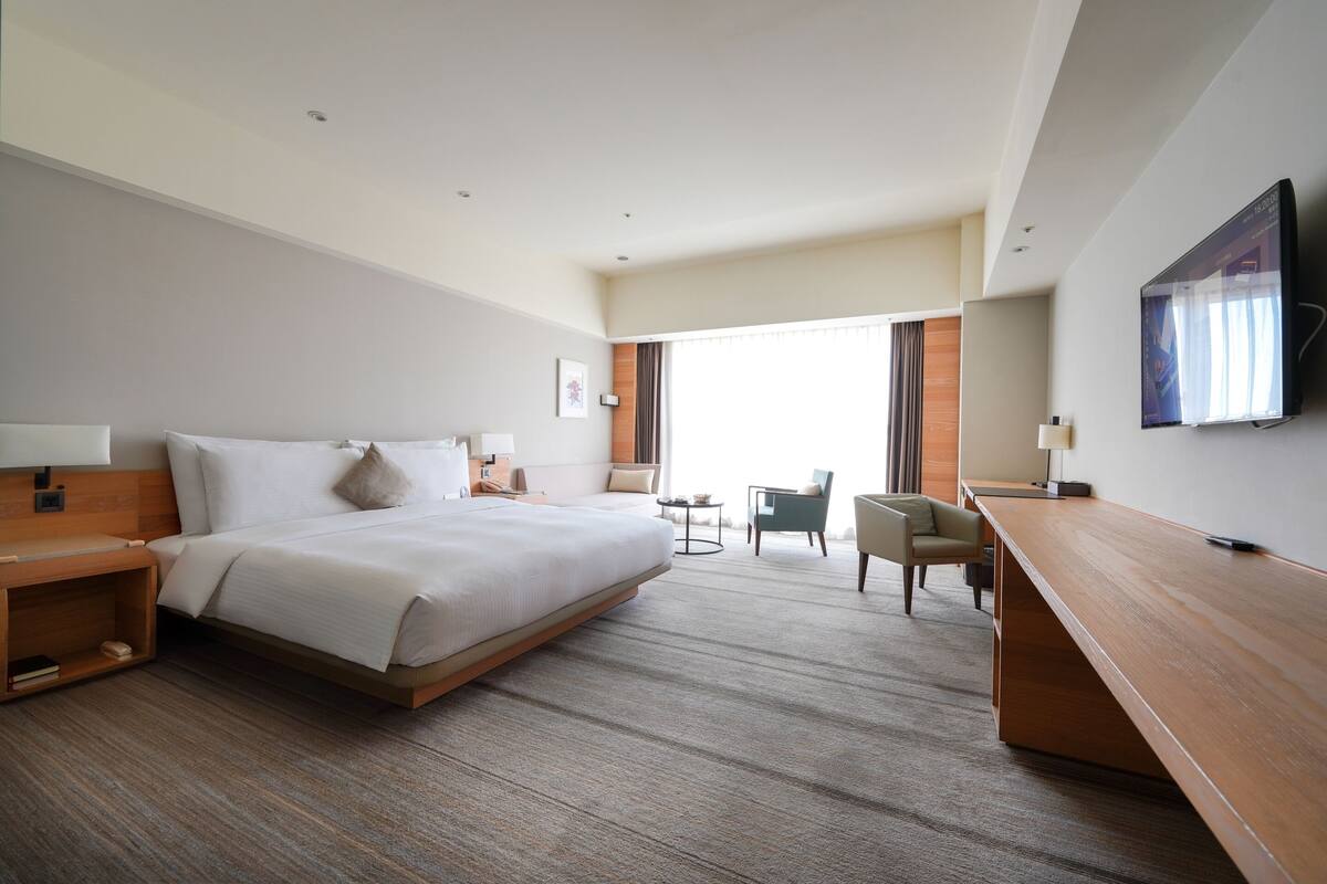 A modern hotel room with a large bed, a desk, a chair, and a TV mounted on the wall.