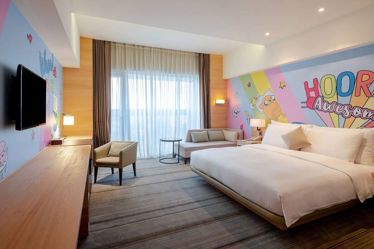 A hotel room with a bed, a chair, a small table, a TV, and a window with curtains.