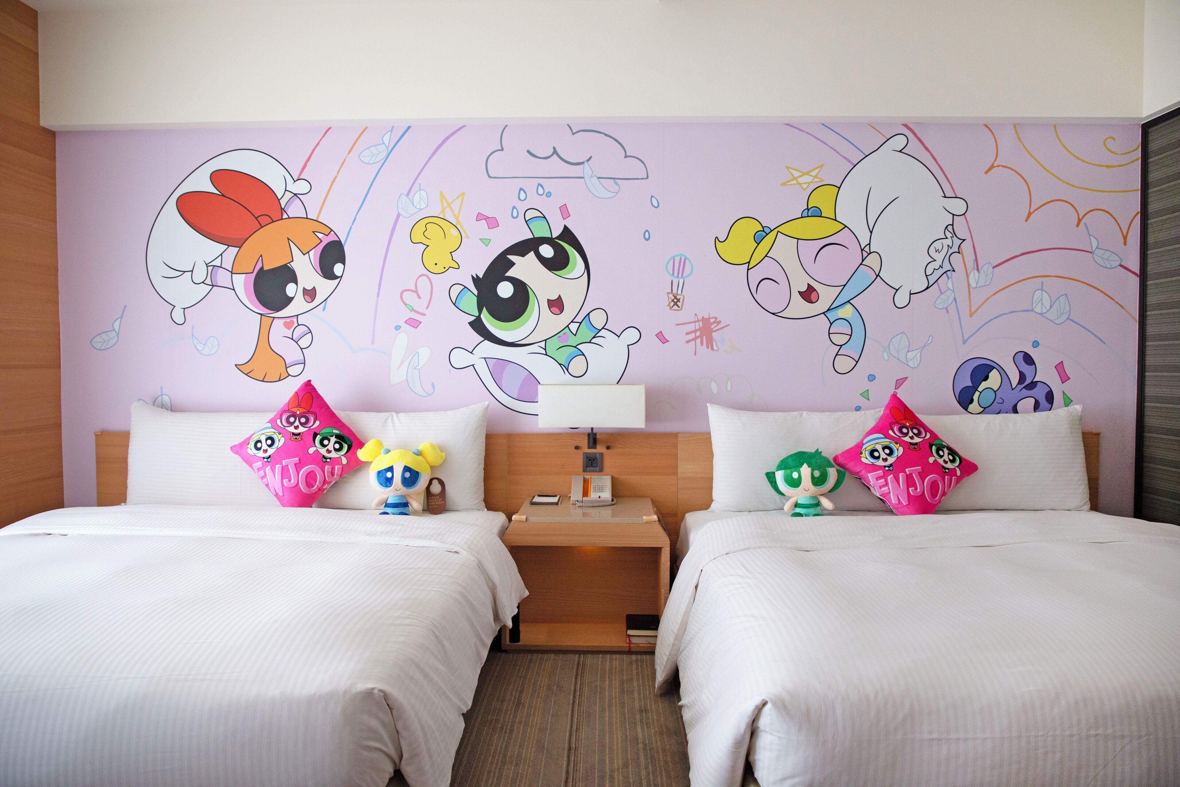 A hotel room with two beds, each with a colorful pillow featuring cartoon characters. There is a nightstand with a lamp and a small clock between the beds. The room has a wooden headboard and a wall with a colorful mural.