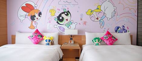 A hotel room with two beds, each with a colorful pillow featuring cartoon characters. There is a nightstand with a lamp and a small clock between the beds. The room has a wooden headboard and a wall with a colorful mural.