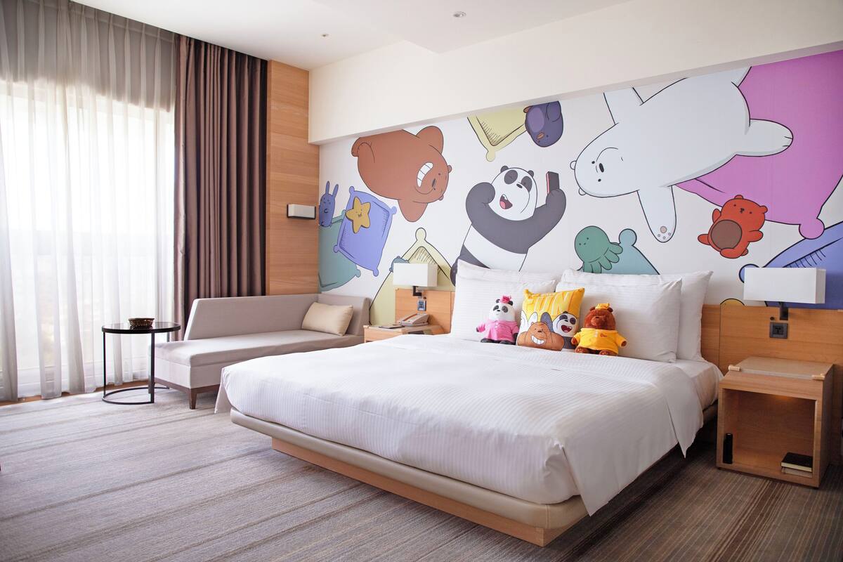 A hotel room with a large bed, a sofa, a small table, and a nightstand. The room has a mural on the wall with cartoon characters.