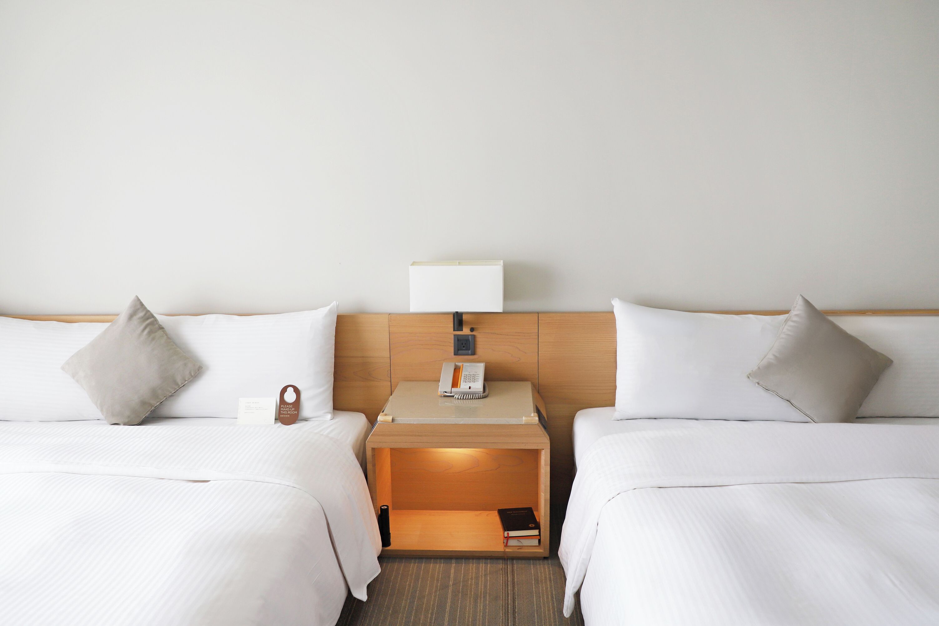 A hotel room with two beds, a bedside table with a lamp, and a small wooden nightstand with a phone and a book.