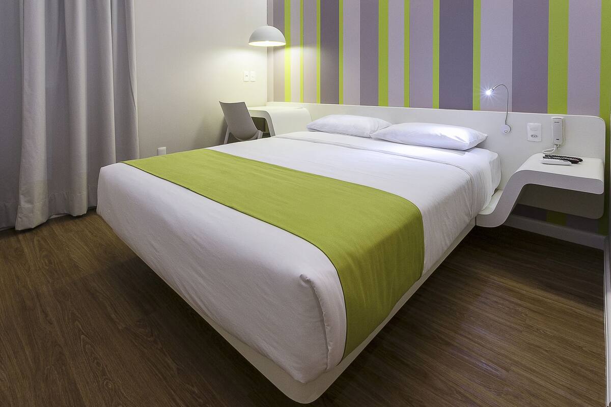 A hotel room with a bed, a bedside table with a lamp, and a wall with vertical stripes.