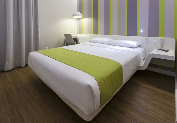 A hotel room with a bed, a bedside table with a lamp, and a wall with vertical stripes. - DMax Hotel (Marilia)