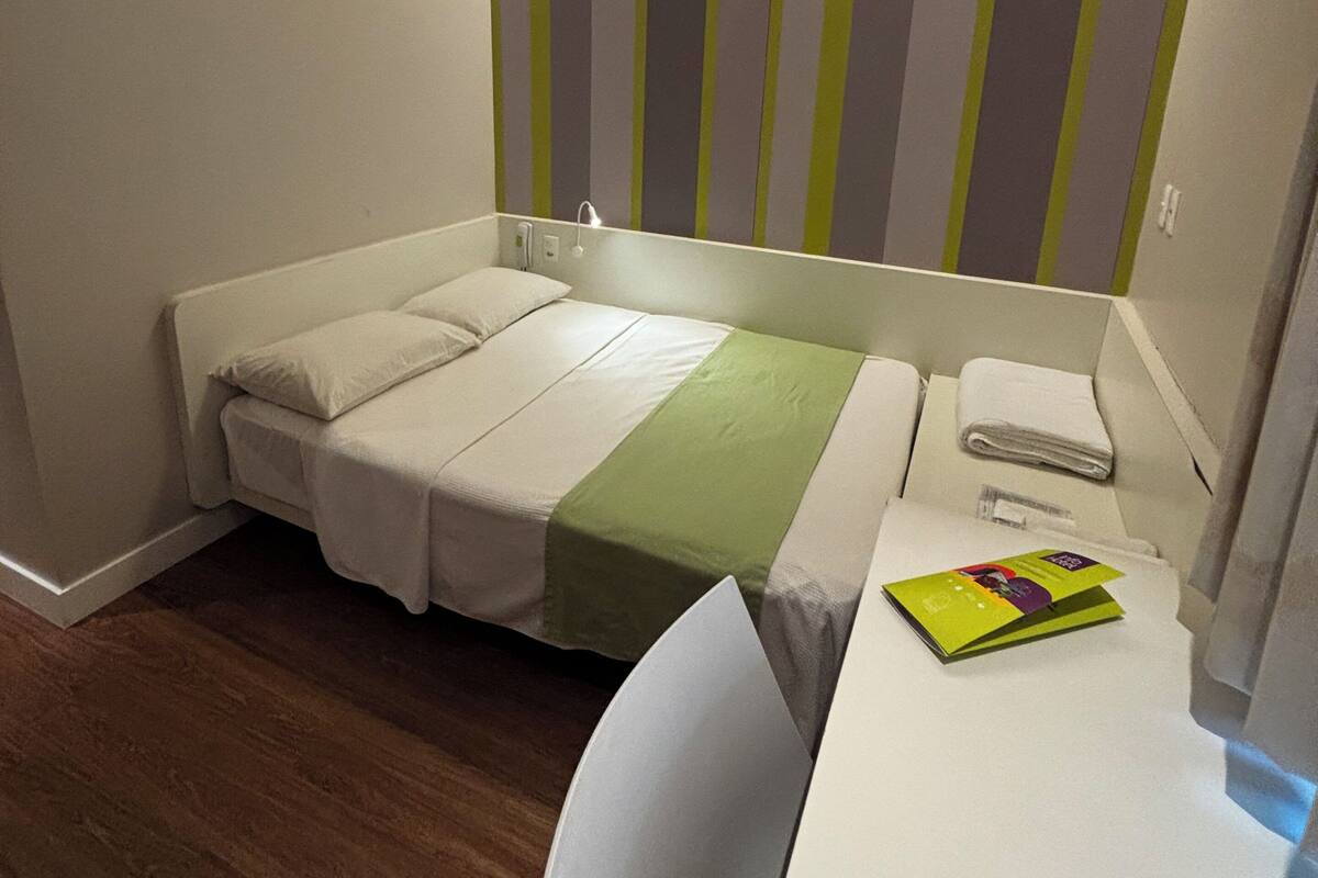 A small, neatly arranged hotel room with a single bed, a desk with a chair, and a striped wall decoration.