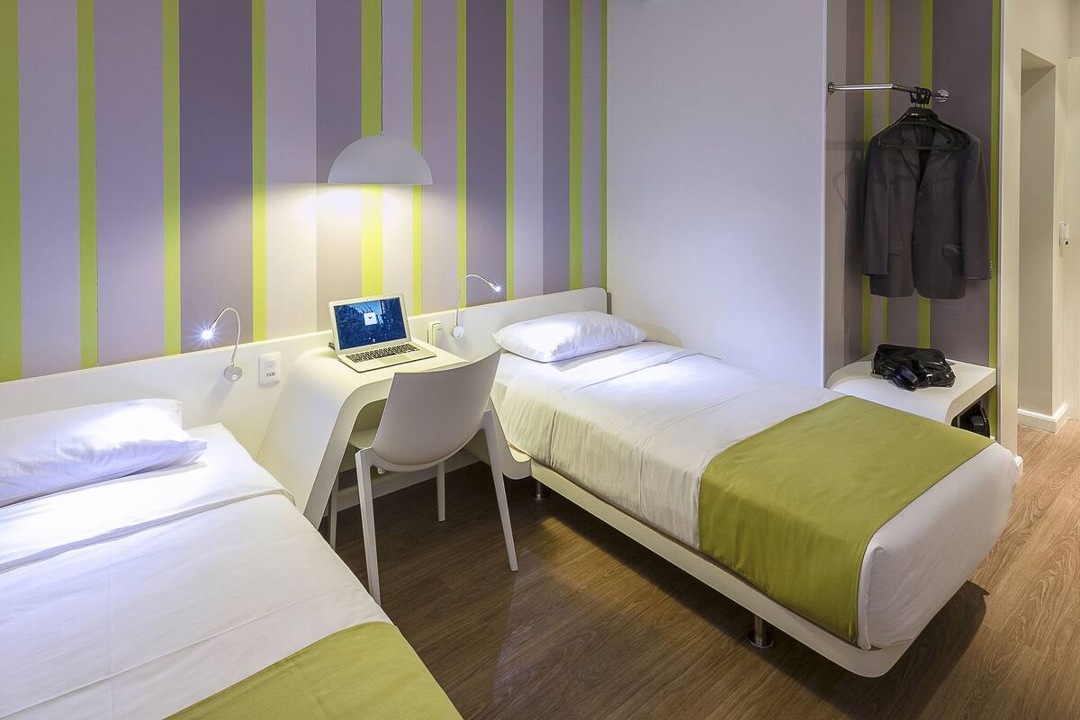 A hotel room with two beds, a desk, a chair, a laptop, a hanging coat, and a bedside table with a bag.