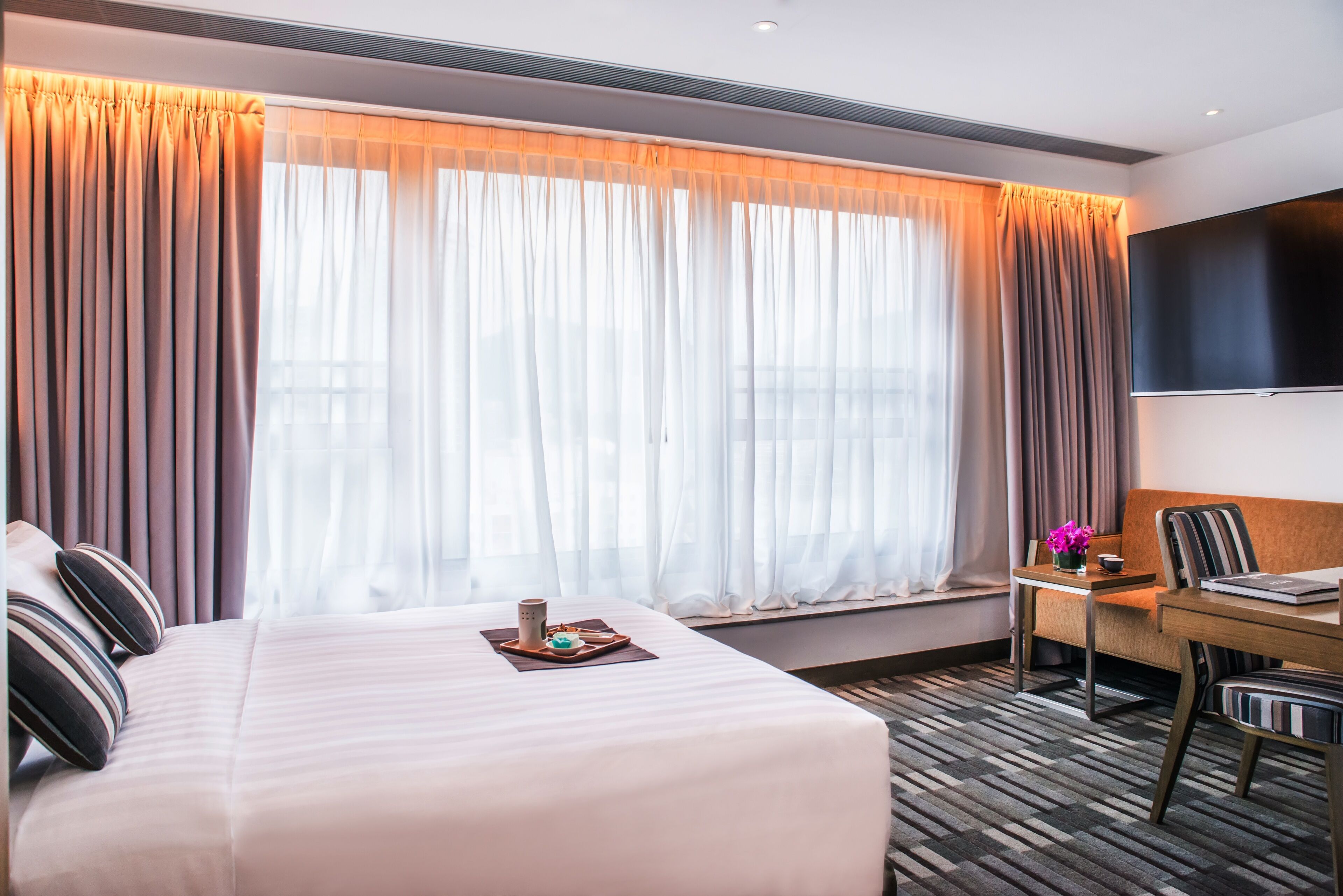 A hotel room with a large bed, a desk with a chair, a flat-screen TV, and a window with curtains.