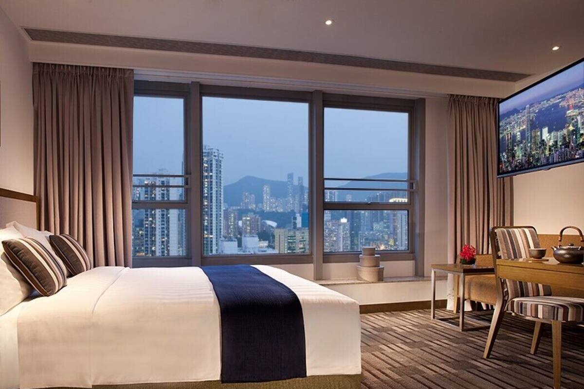 A hotel room with a large bed, a dining table, and a city view through large windows.