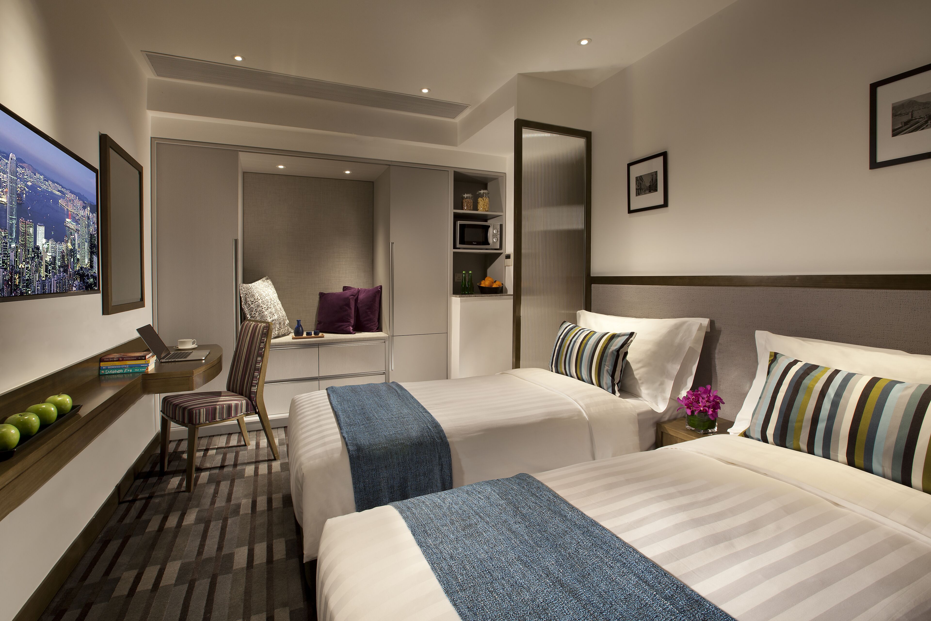 A hotel room with two beds, a desk with a laptop, a chair, a bookshelf, and a cityscape painting.