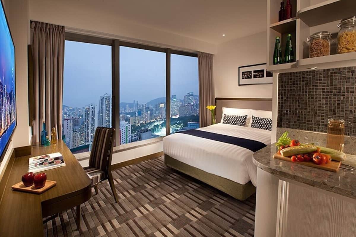 A hotel room with a large window, a bed, a desk, and a kitchenette.