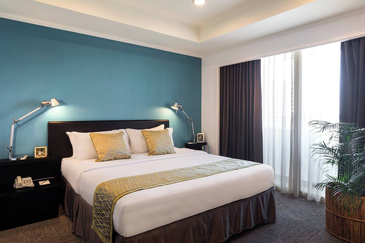 A hotel room with a large bed, two bedside lamps, a nightstand, a telephone, and a potted plant.