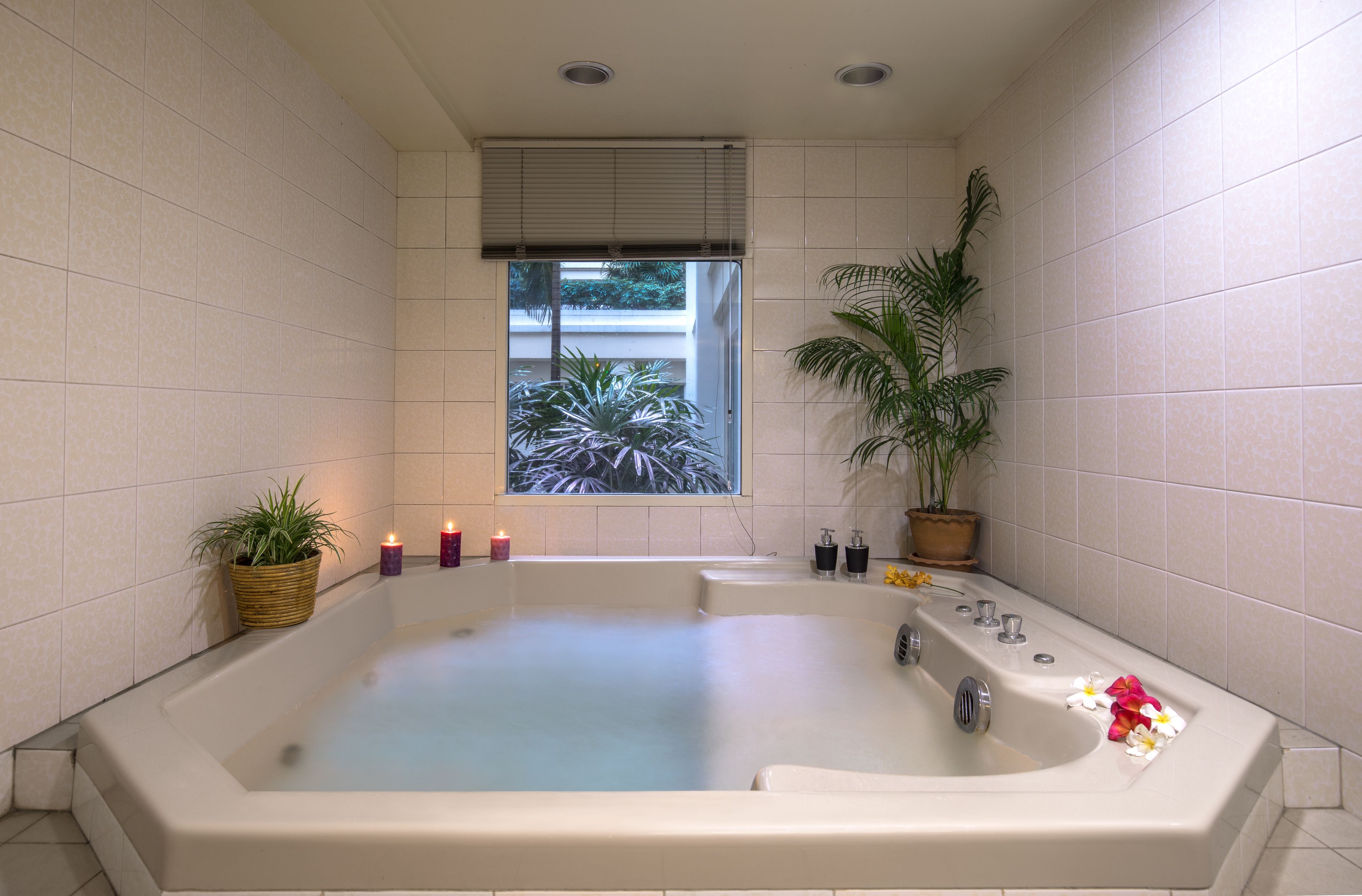 A large, rectangular bathtub with a window above it, surrounded by white tiled walls and a potted plant.