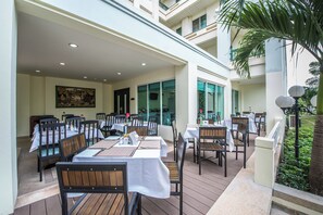 Un área de comedor al aire libre con varias mesas y sillas, una terraza de madera y vistas a un edificio con balcones.