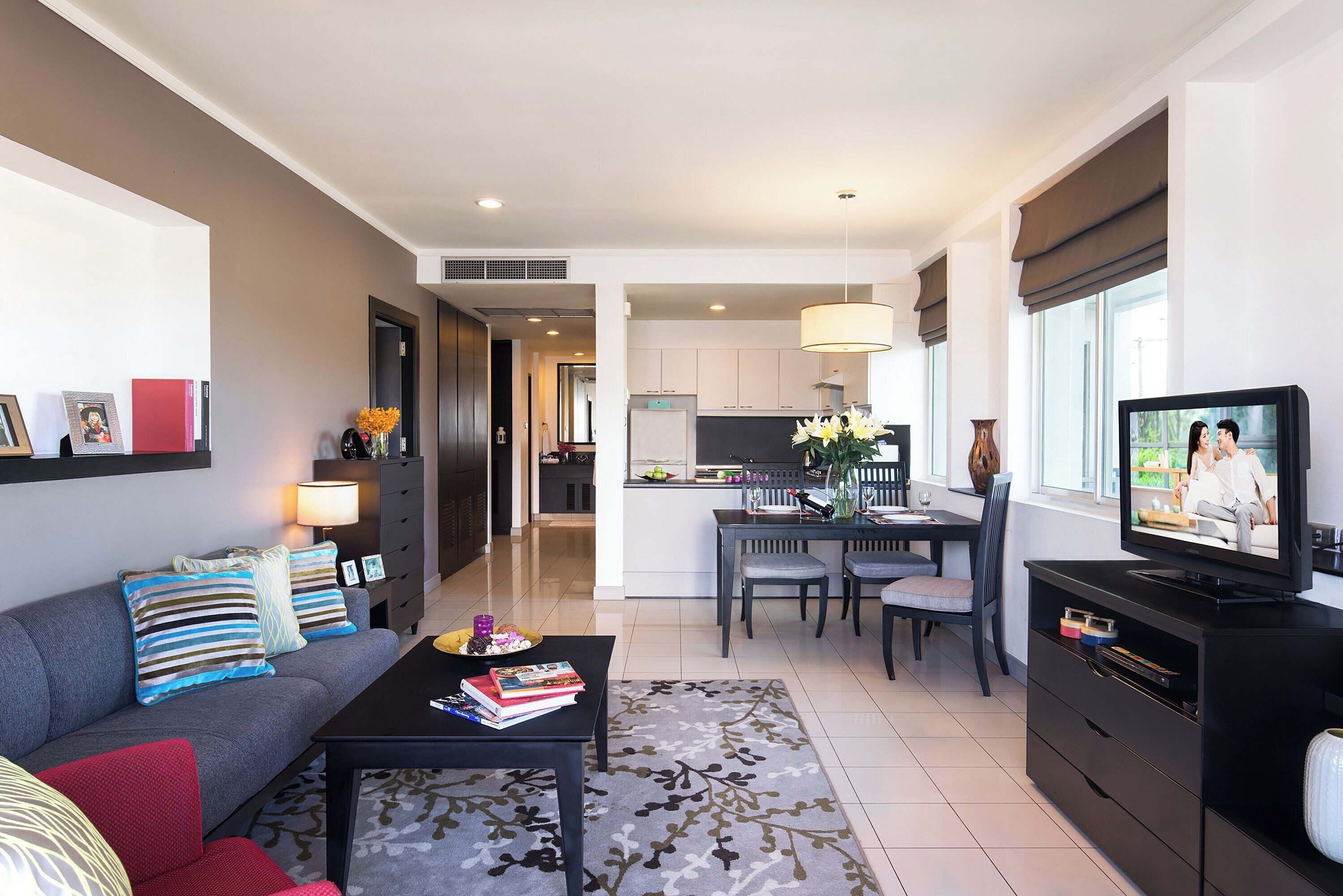 A modern living room with a sofa, coffee table, and TV. There is a dining area with a table and chairs, and a kitchen in the background.
