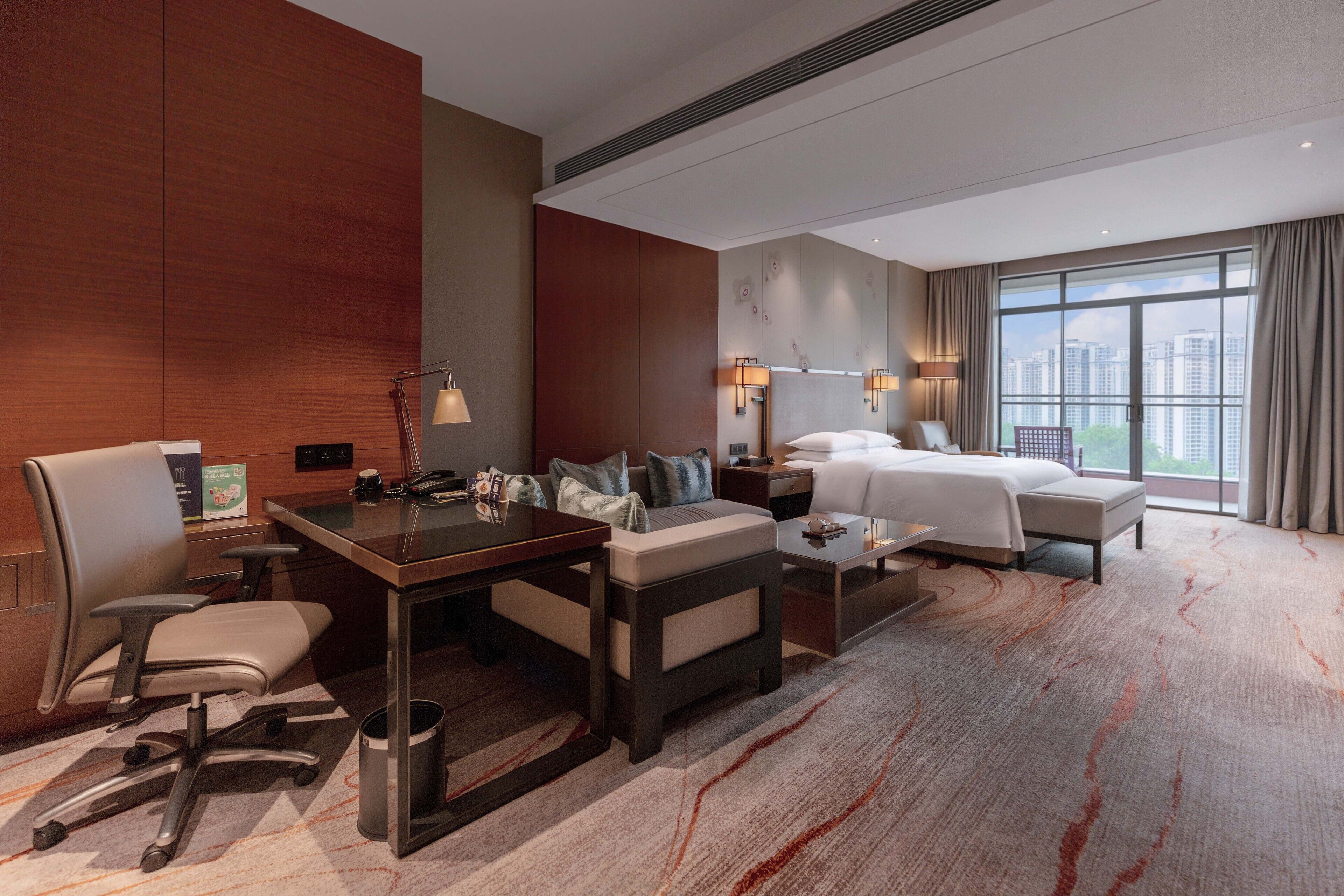 Habitación de hotel con una cama grande, un escritorio con una silla, un sofá y una mesita pequeña. Hay dos lámparas de pared, una ventana con cortinas y vistas a edificios.