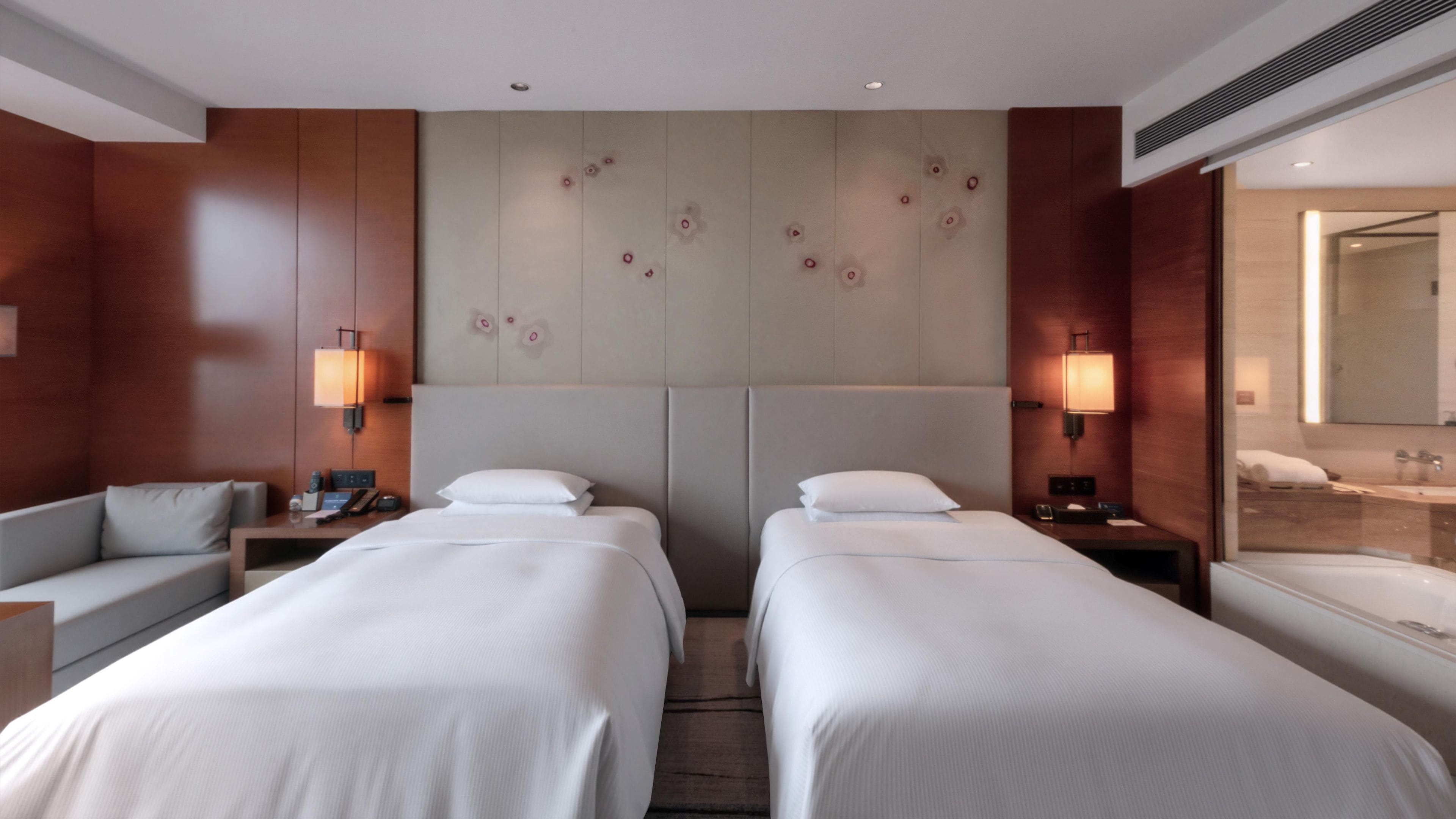 A hotel room with two beds, a sofa, a desk, a lamp, a mirror, and a bathroom visible through the door.