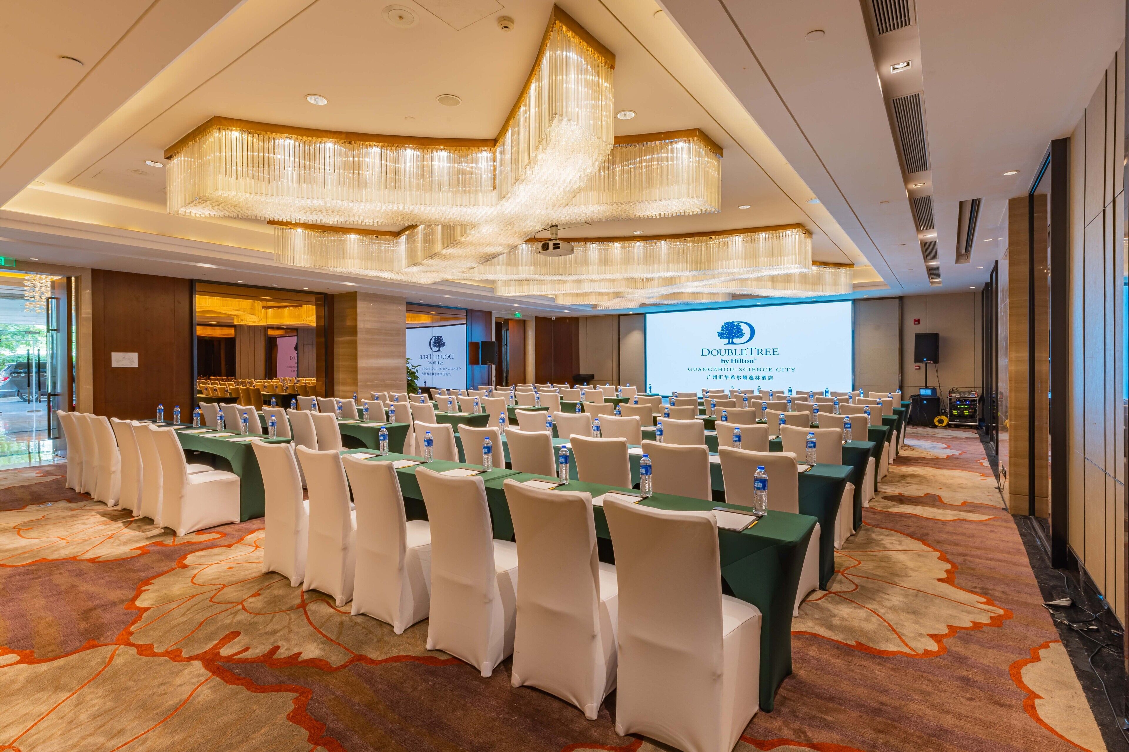 A conference room with rows of tables and chairs, a large screen, and a chandelier.