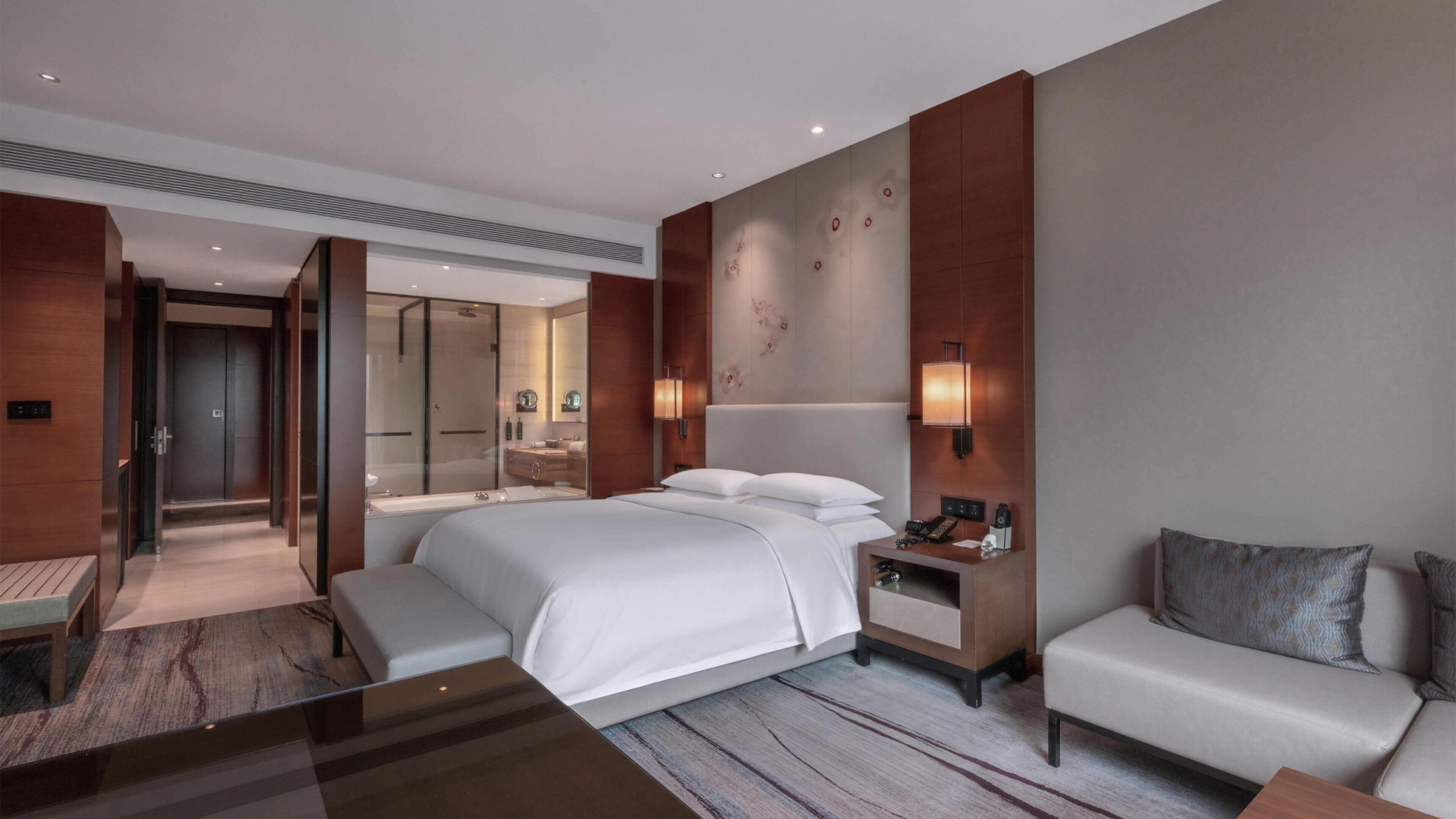 A modern hotel room with a large bed, bedside table, lamp, and a sofa.