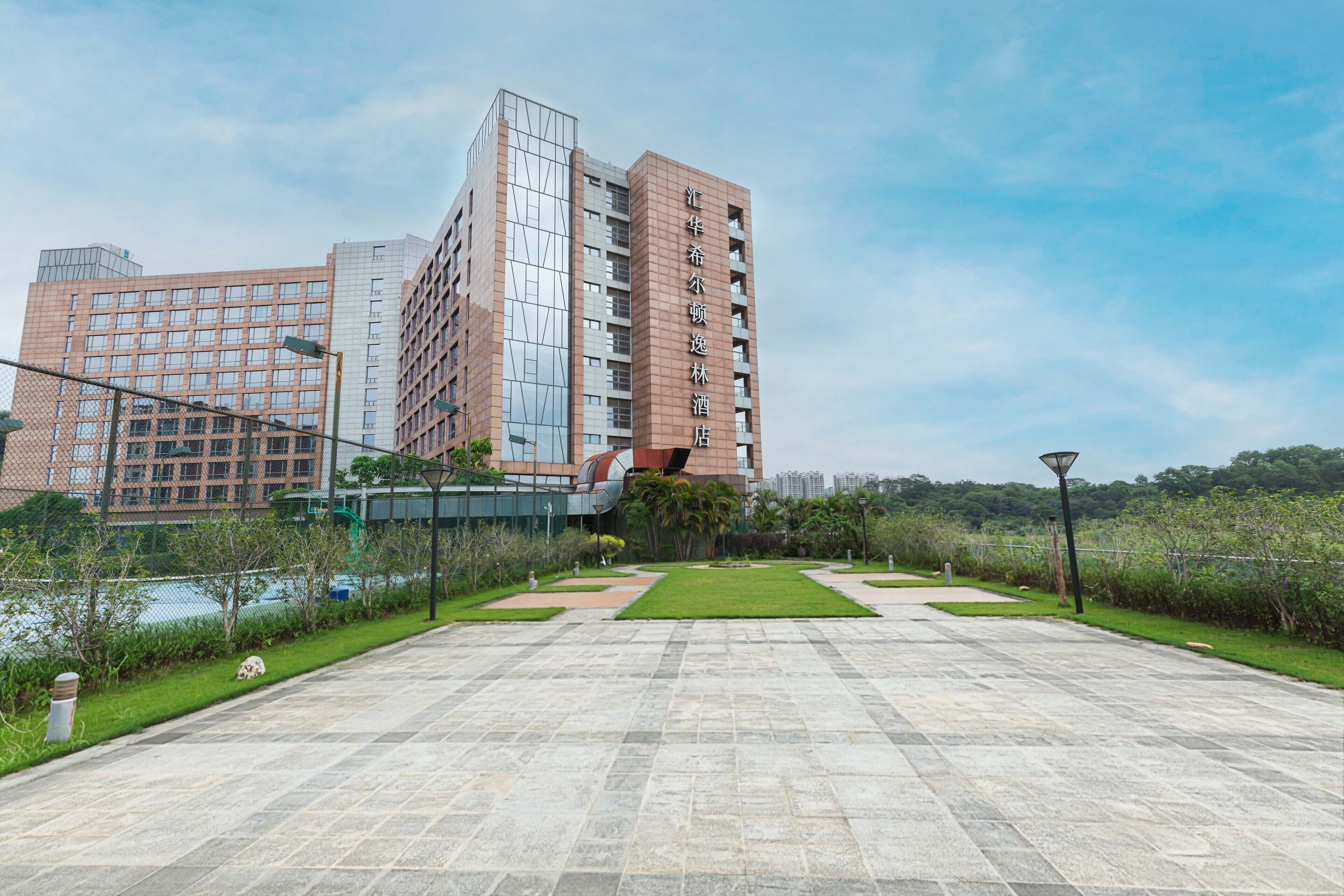 A modern hotel with a prominent glass facade, a well-maintained courtyard with pathways, and a view of a river.