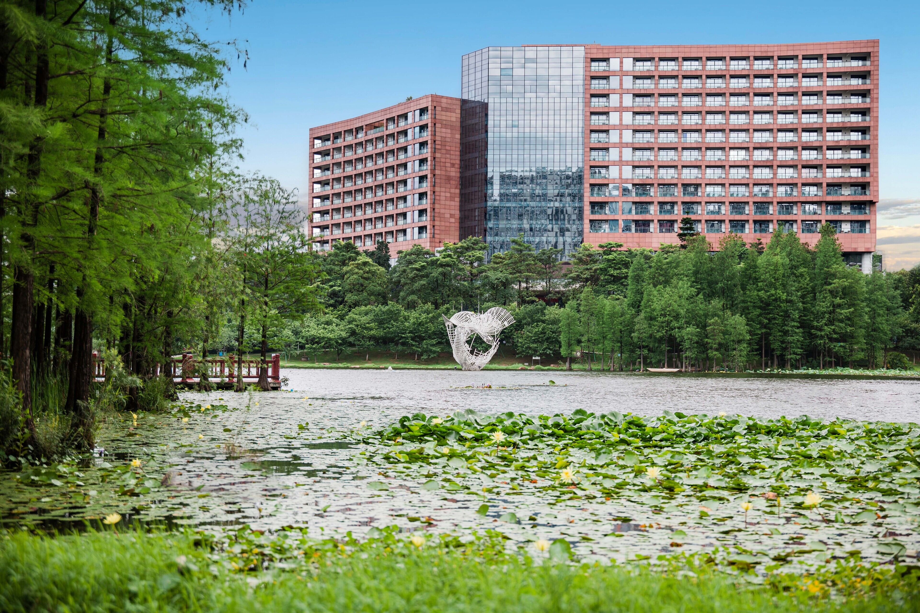 Um parque com um lago, rodeado por árvores e uma escultura em forma de coração. Ao fundo, dois edifícios altos, um deles com fachada de vidro.
