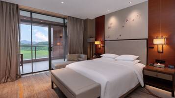 A modern hotel room with a large bed, a bedside table with a lamp, a nightstand, and a view of a balcony with a glass door.