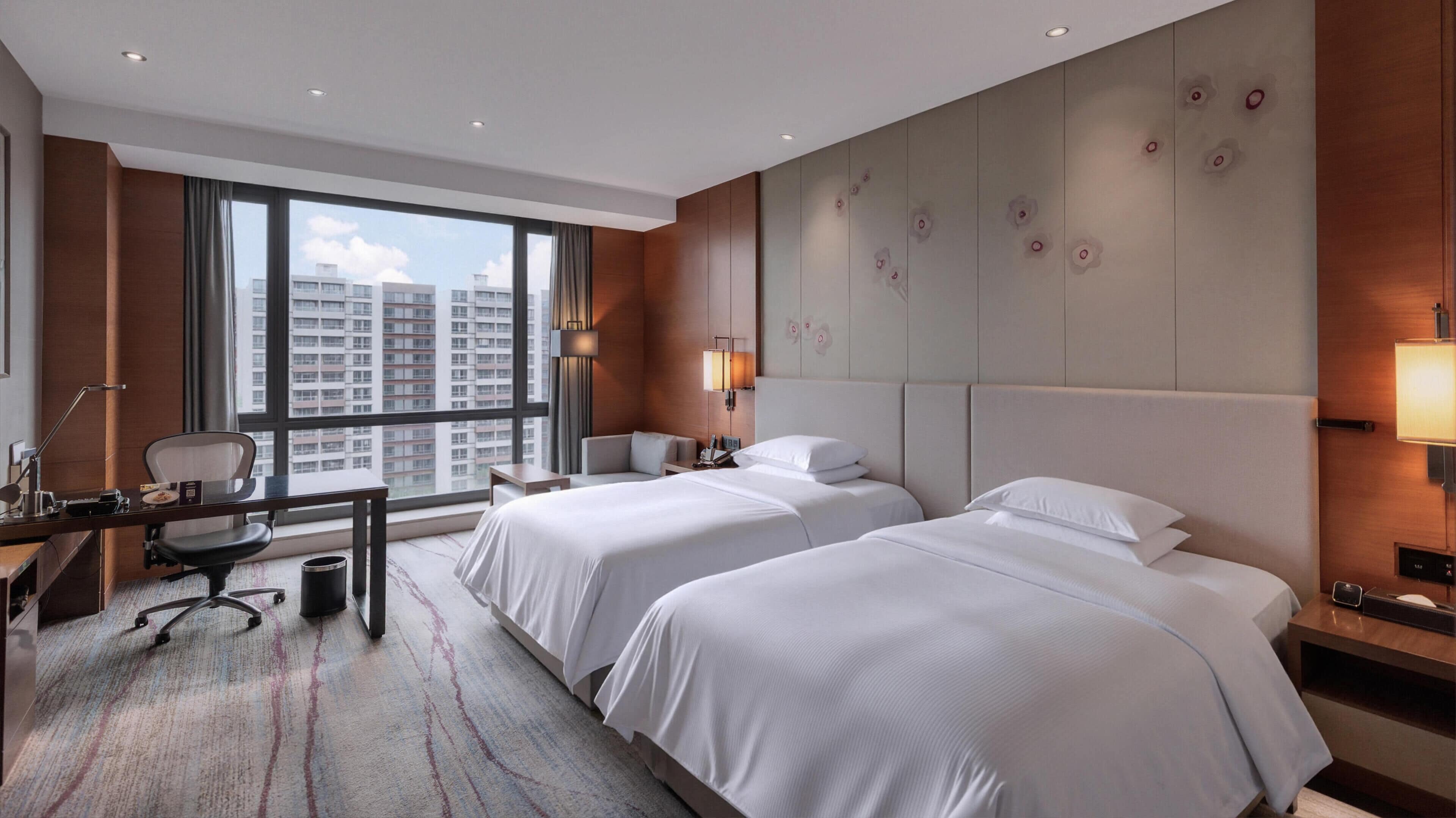 Une chambre d’hôtel avec deux lits, un bureau, une chaise, une lampe et une grande fenêtre donnant sur la ville.