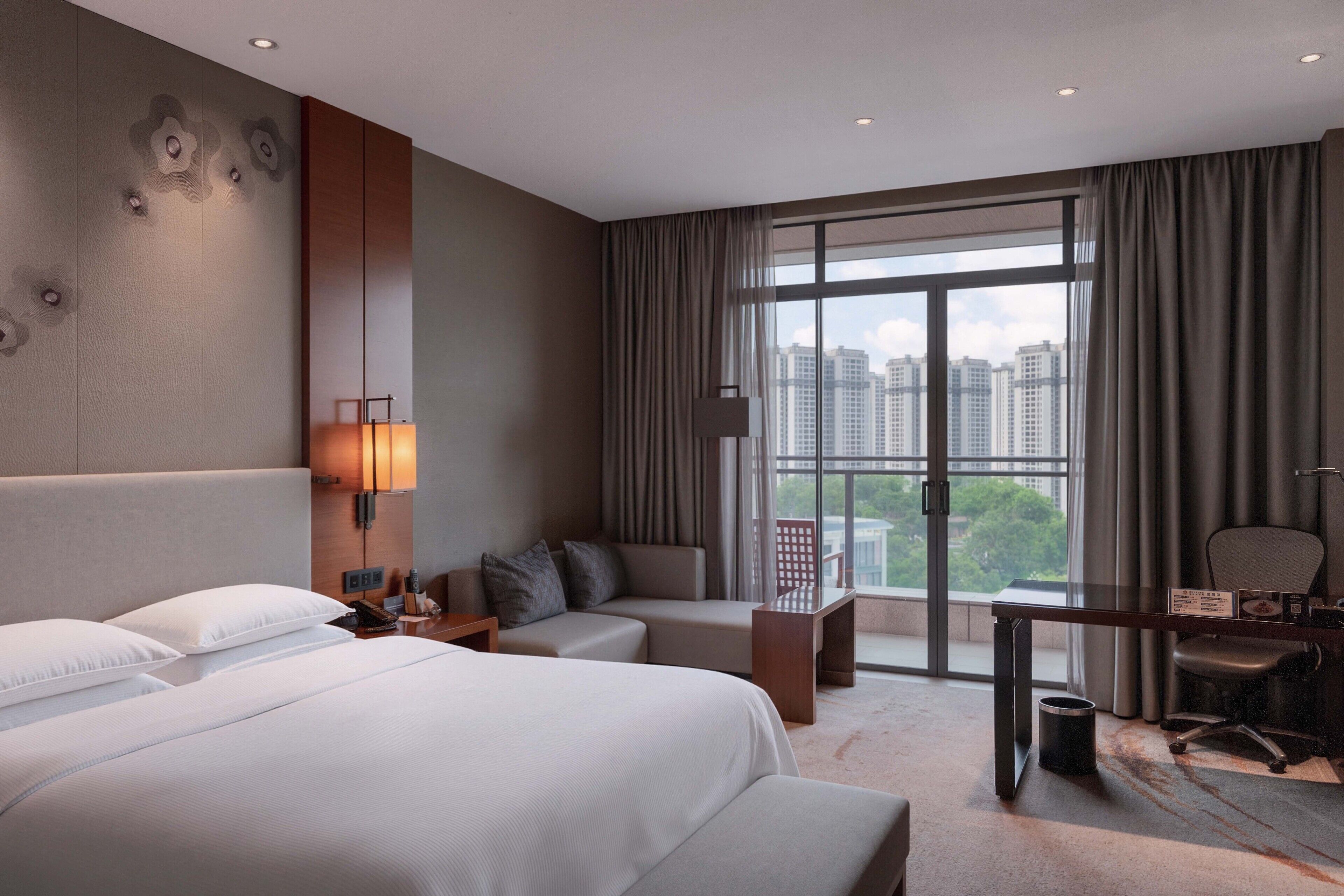 A hotel room with a large bed, a sofa, a desk, and a view of the city through large windows.