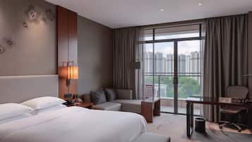 A hotel room with a large bed, a sofa, a desk, and a view of the city through large windows.