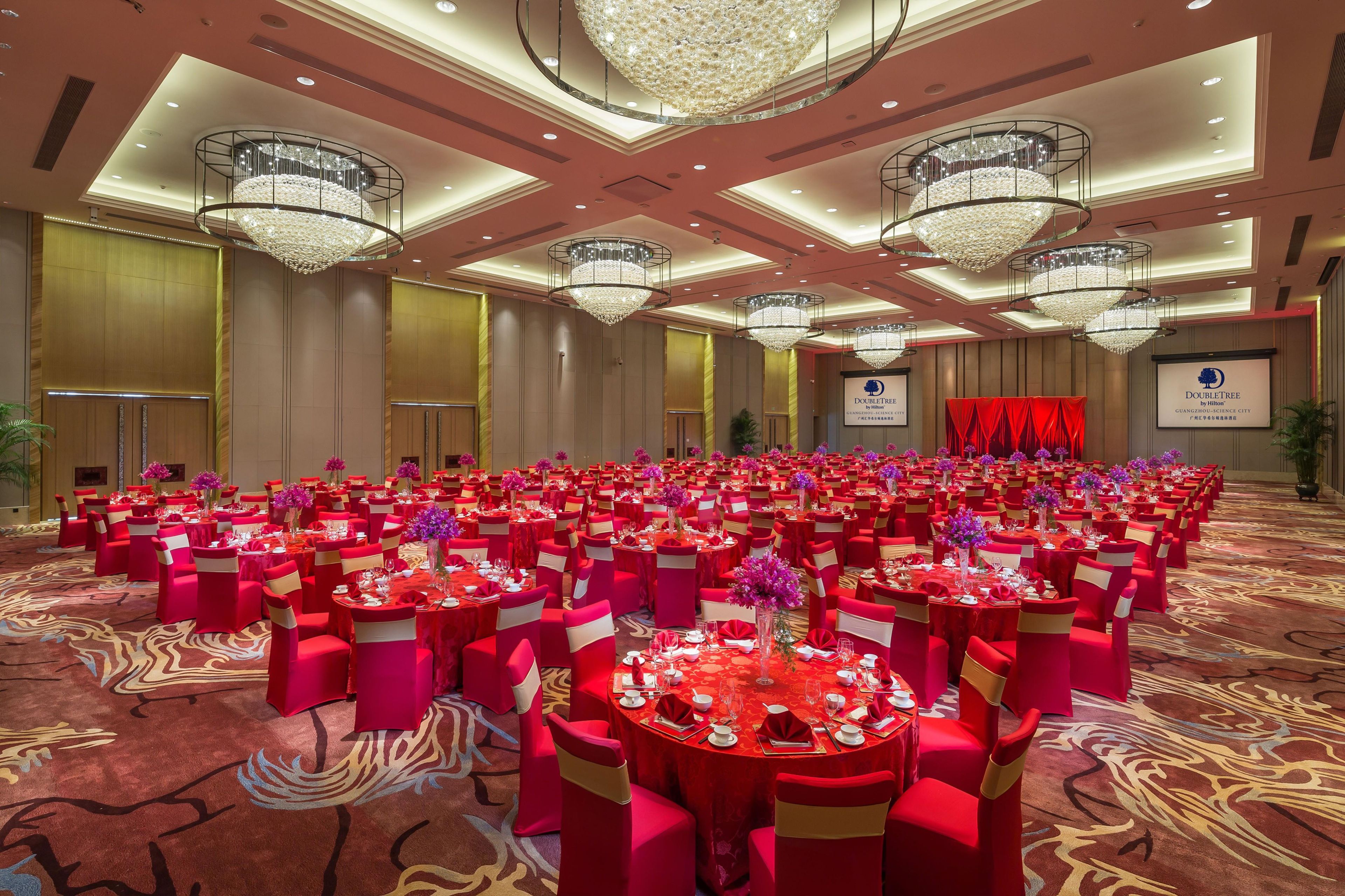 A banquet hall with round tables, red chairs, and chandeliers.