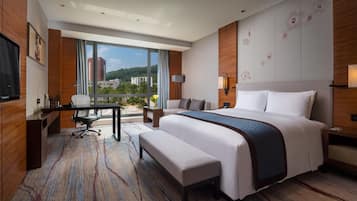 Habitación de hotel con una cama grande, un escritorio, una silla, un banco, un televisor y vistas a edificios a través de amplias ventanas.