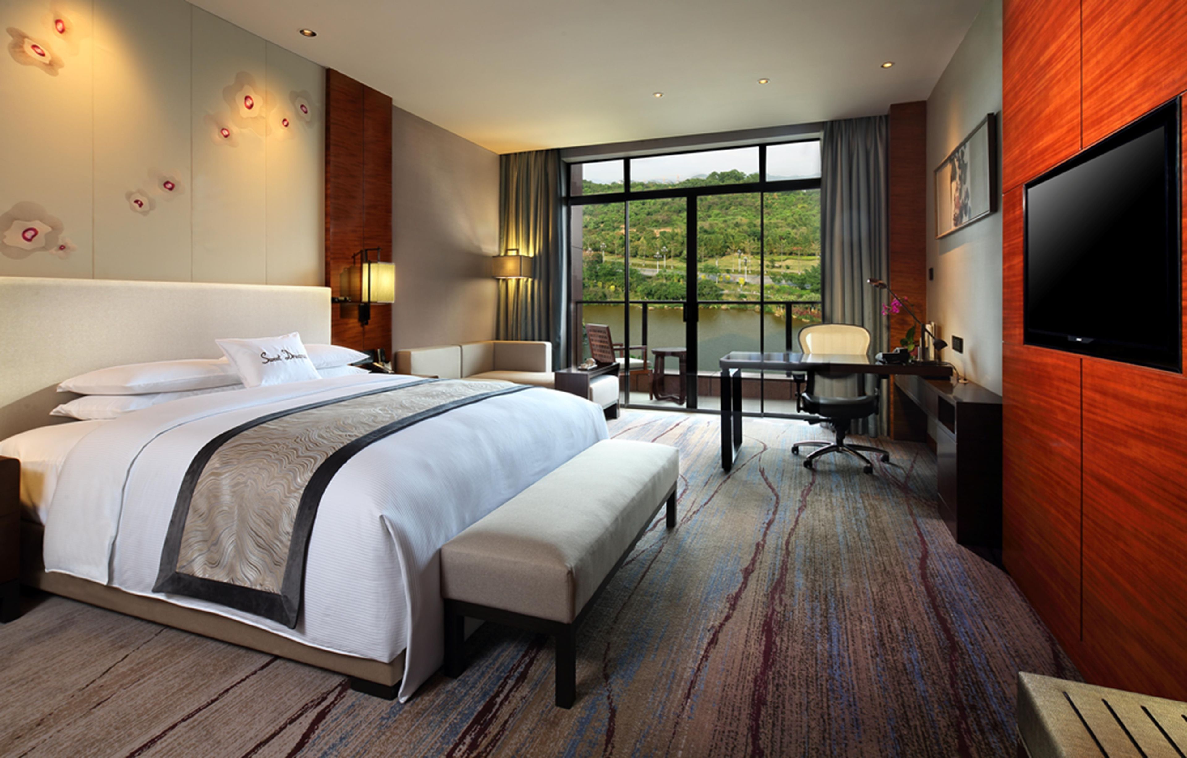 A hotel room with a large bed, a desk with a chair, a TV, and a view of a lake.